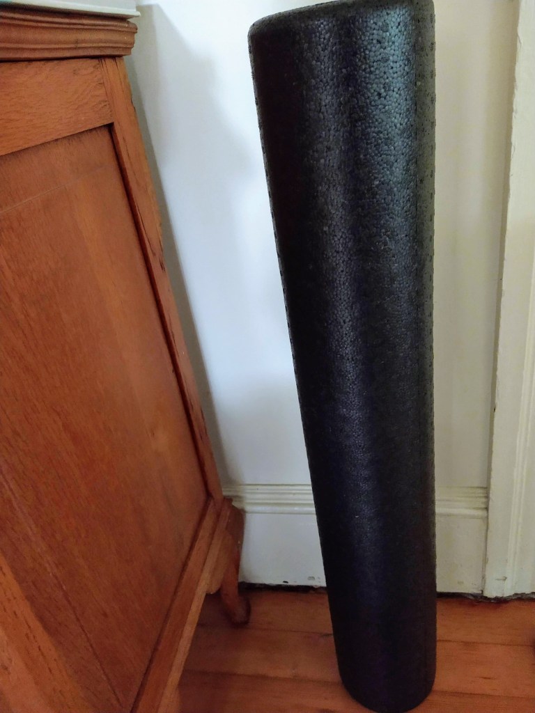 A black foam roller I use for back and leg massages. Upright in a living room near furniture.