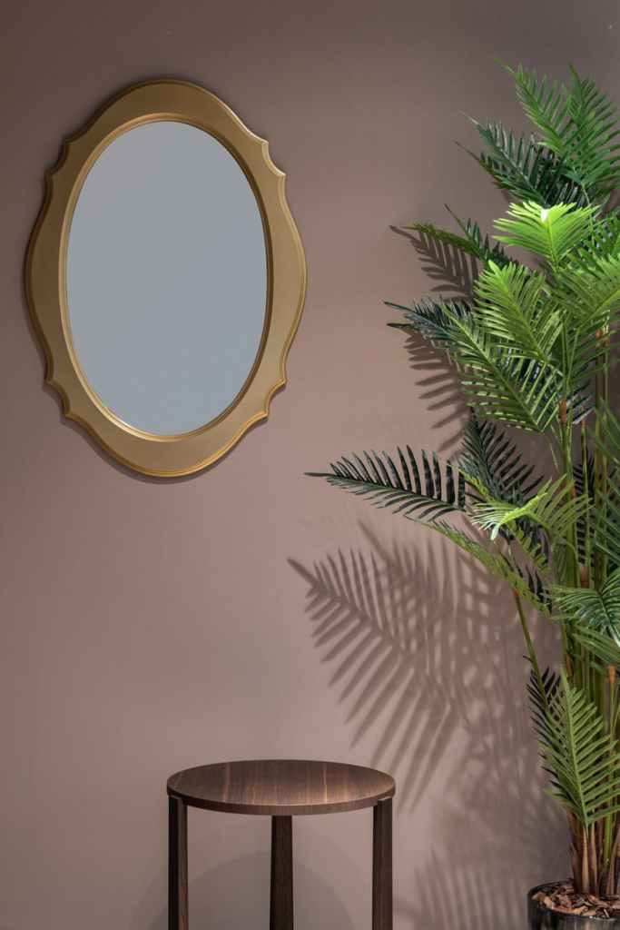 Mirror with gold edges on a dark pinkish wall. Fern Plant in a vase, mahogany stool.