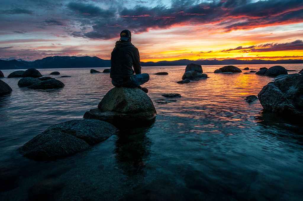 Man sitting on a rock near the shore of an ocean, staring at the sunset.