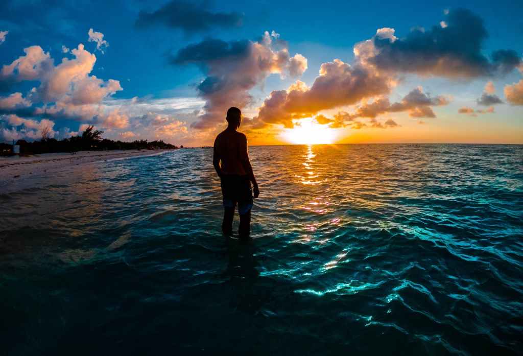 Shadowed Man standing in turquoise waves staring at a setting sun in a tropical place.