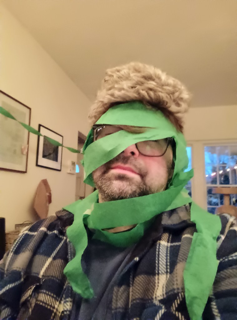 Selfie of a man wrapped in party decorations with a wig on, sitting down. White walls behind him.