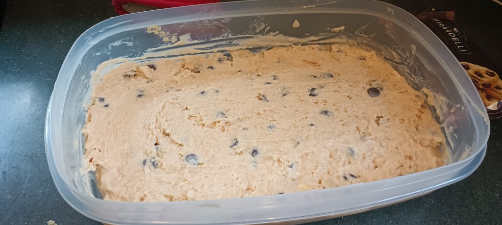 Picture of the dairy free chocolate chip icecream in a 12 cup plastic tupperware container. Container is on a green tile counter with ghiradelli chocolate chip package in the top right corner.