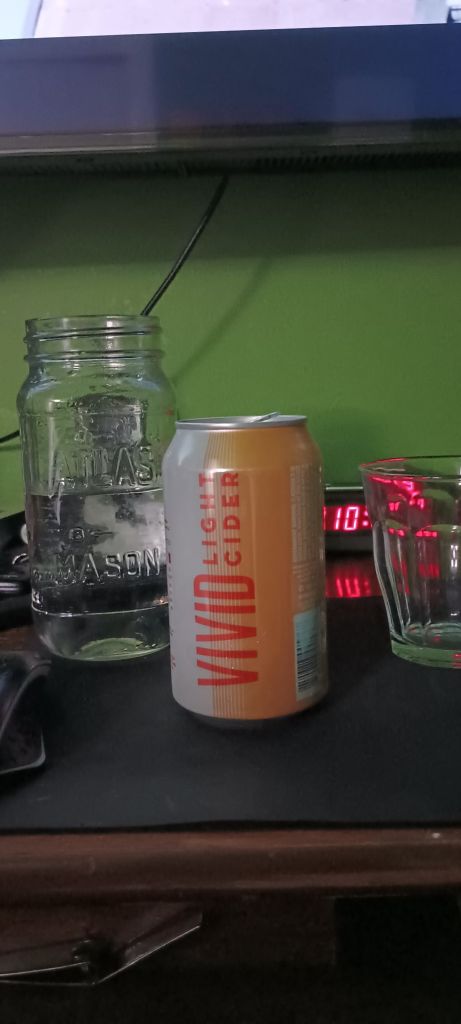 Glass of water, a 12-oz can of Seattle Cider Vivid light cider, peach flavor. With an empty glass, a green background, and an alarm clock with red text displaying 10 pm and unreadable, distorted integer.